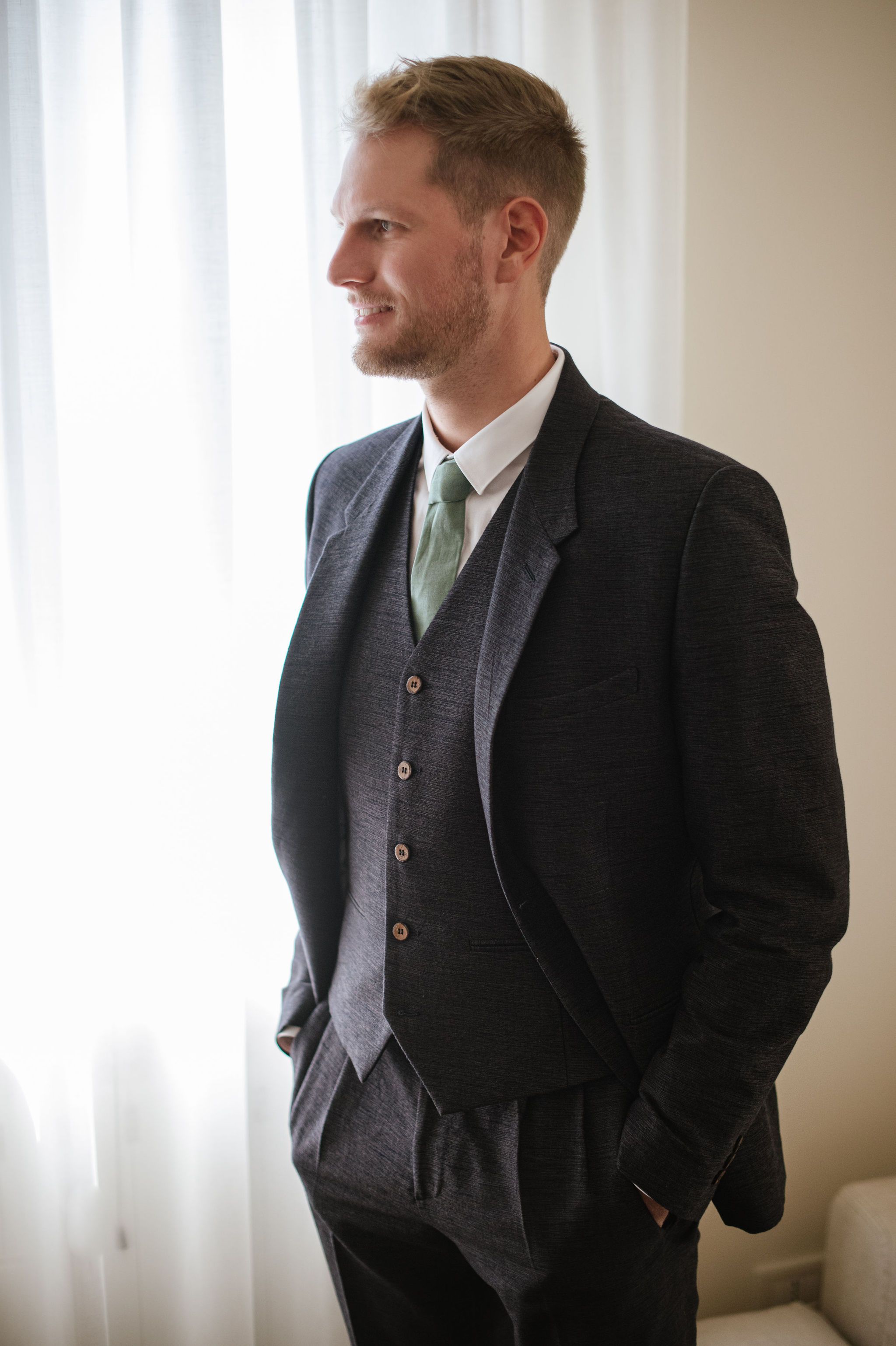 Groom in dark three-piece suit with mint green tie looking out window.