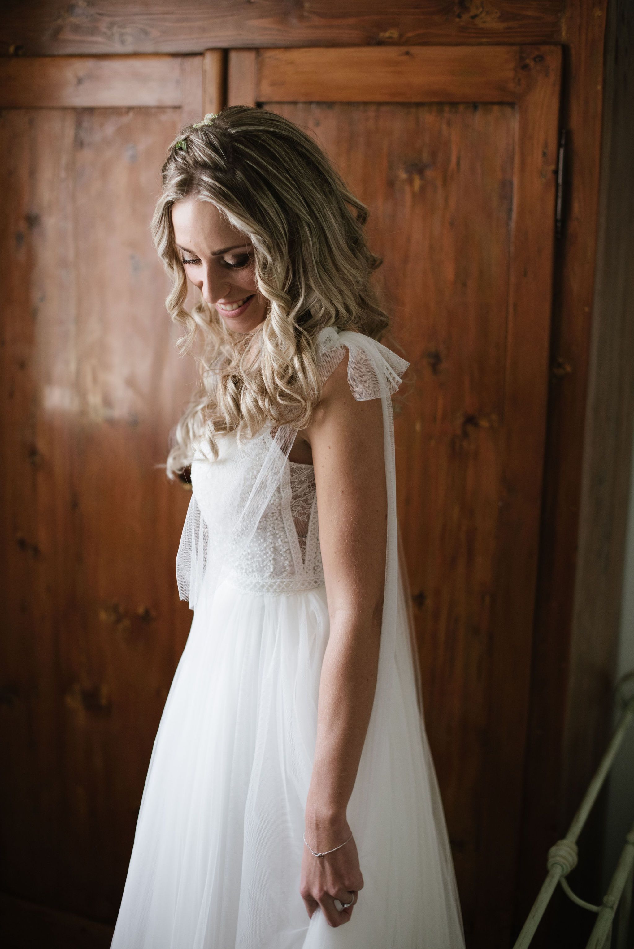 Bride in flowing white dress with lace details smiling against wooden door.