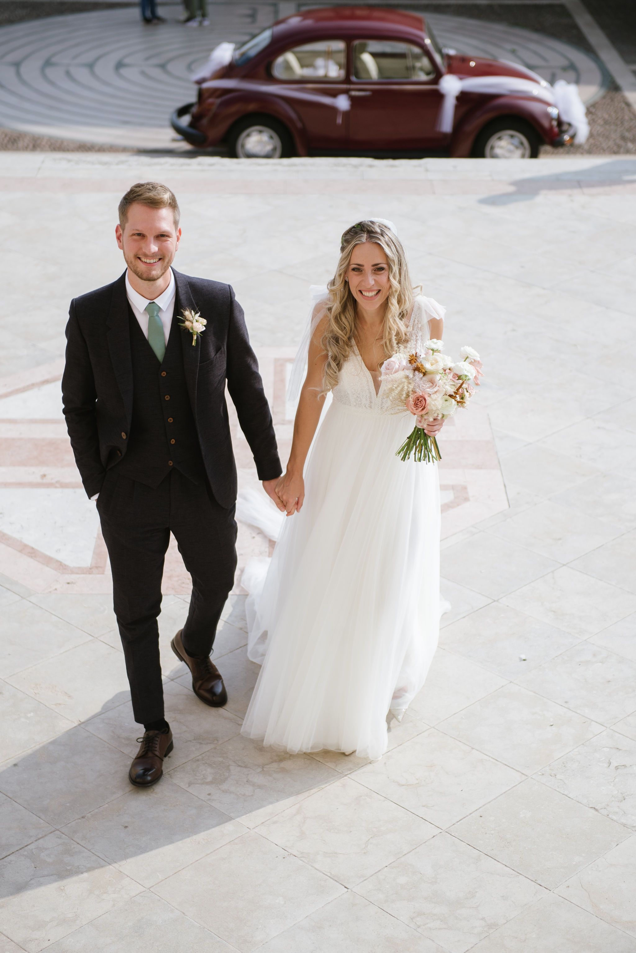 Wedding couple walking hand in hand with vintage red Volkswagen Beetle in background.
