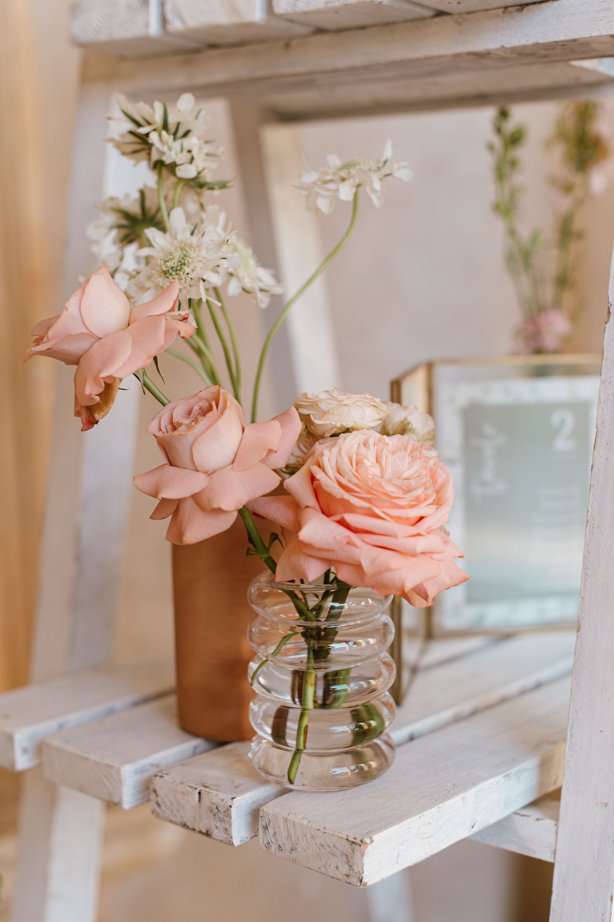 Delicate pink roses and white flowers arranged in clear glass jar on white shelf.