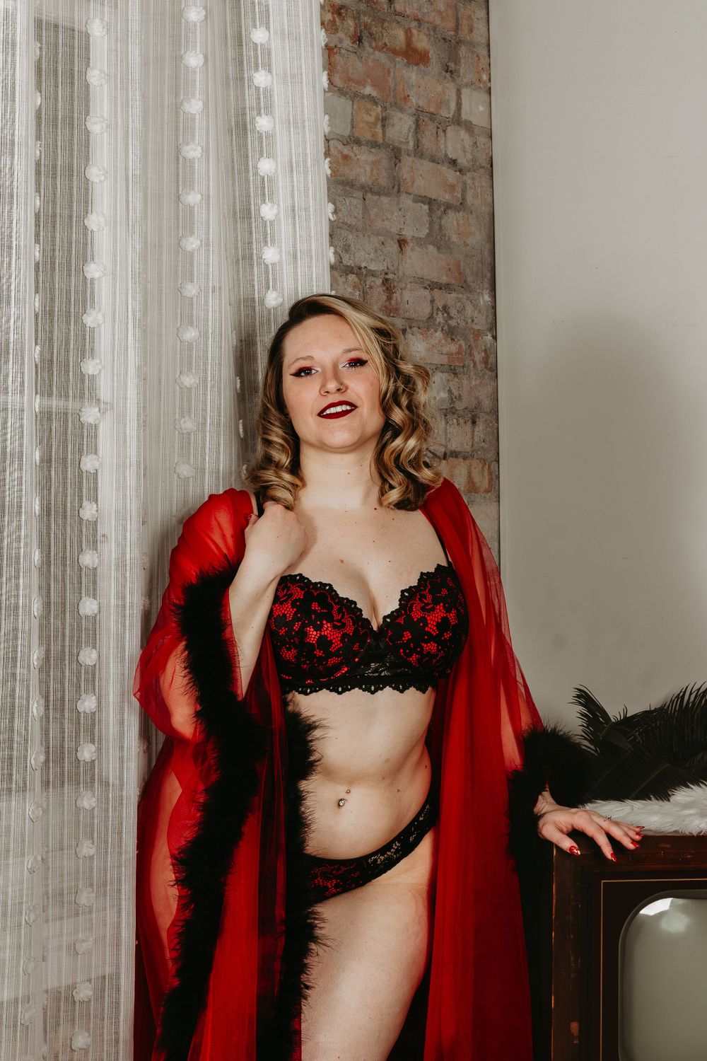 Boudoir photo in vintage style with red sheer robe and black lace lingerie against exposed brick wall and sheer curtains.