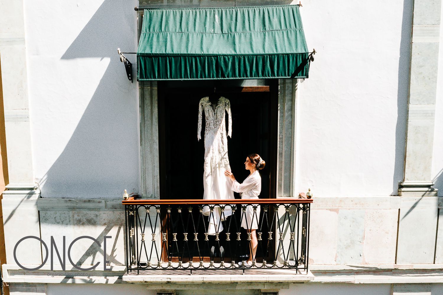 A wedding dress hangs on display under a green awning on a white building's balcony.