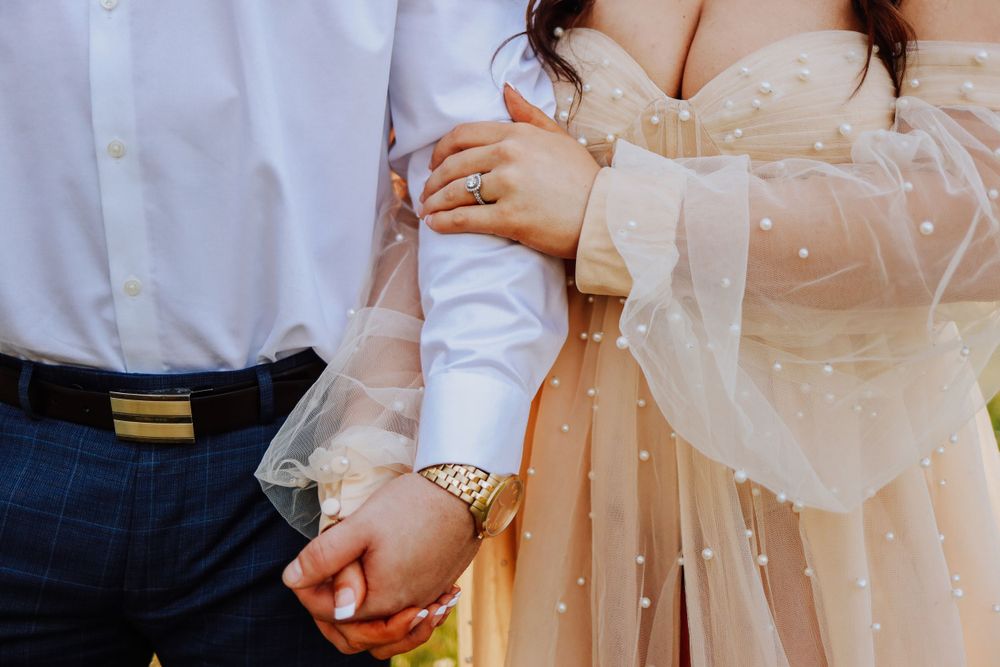 Close-up of an engaged couple holding hands, woman wearing a pearl-embellished champagne dress and ring.