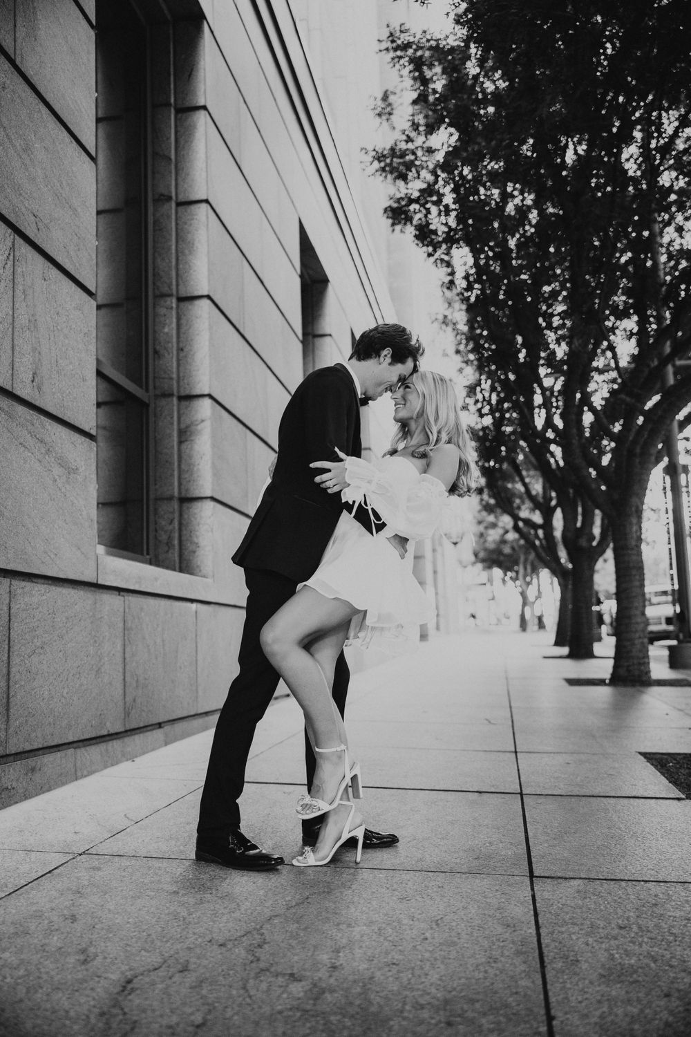 Black and white photo of couple sharing a dip kiss on urban sidewalk, tuxedo and white mini dress.