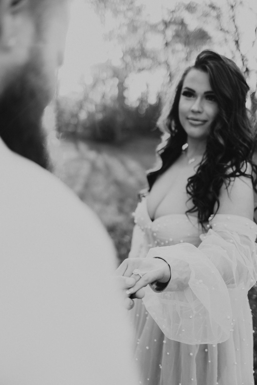 Black and white photo of a brunette woman in a sheer gown holding hands with her partner outdoors.