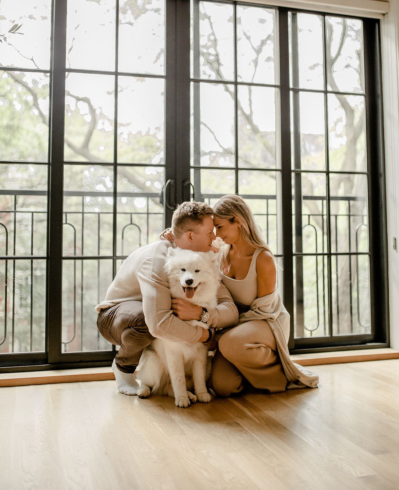 Couple cuddling with white Samoyed dog by large windows in sunlit room.