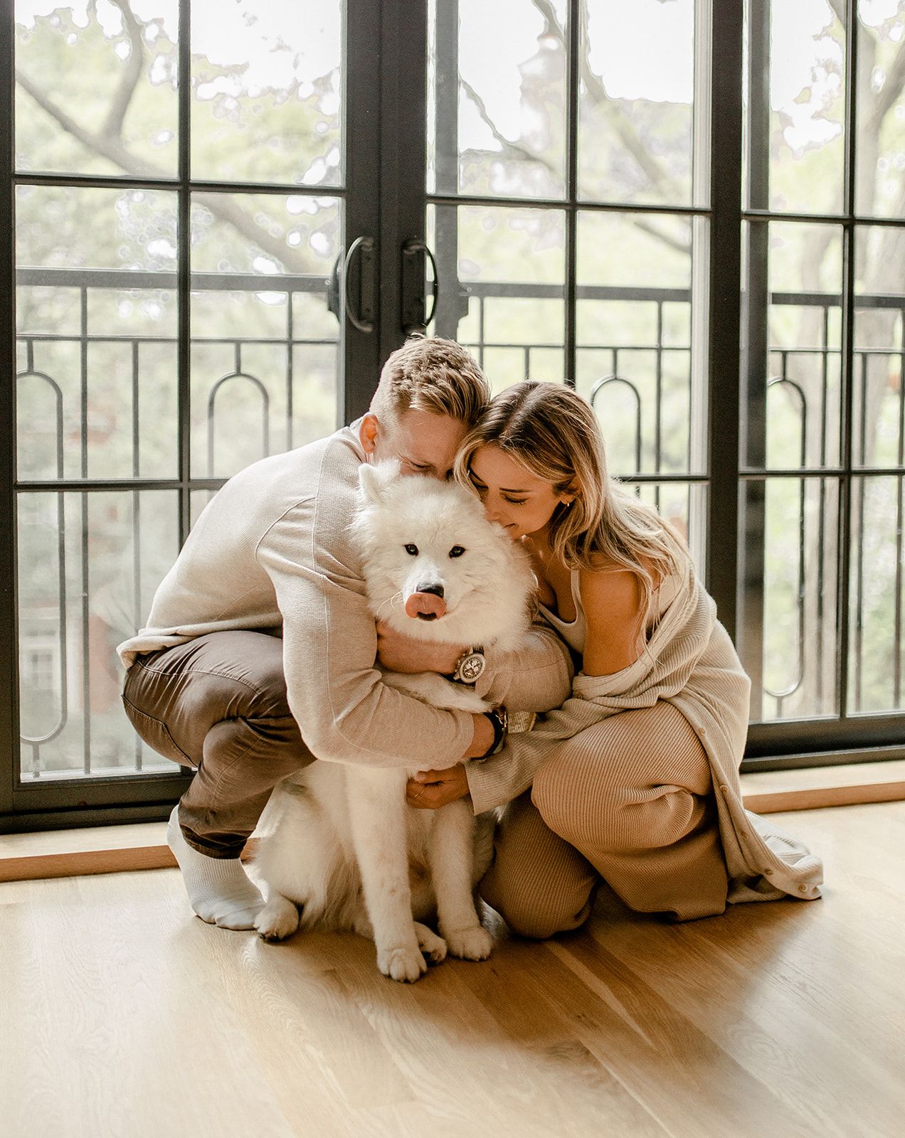 Cozy indoor portrait of couple embracing their fluffy white dog by sunny windows.