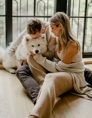 A cozy indoor photo with a couple and their white Samoyed dog by large windows.