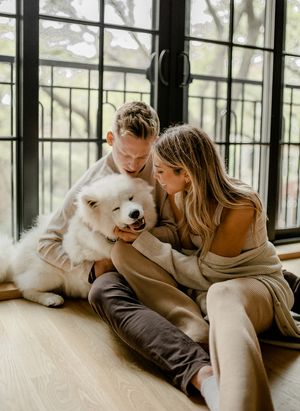 A loving couple cuddles with their smiling Samoyed dog near bright windows.
