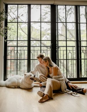 A couple sits with their white fluffy dog by large windows in a modern apartment.