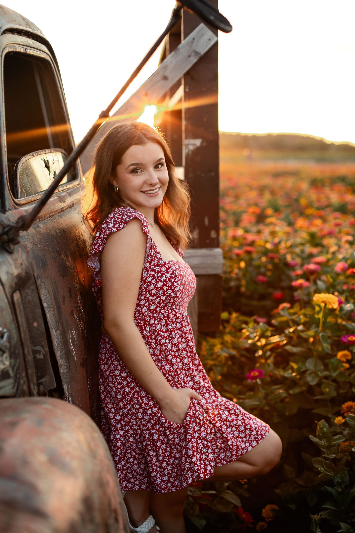 Smiling teenage girl in floral dress leaning against vintage truck in a colorful flower field at sunset.