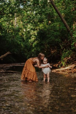 A big sister in an orange dress, leaned over kissing her sister on the head, they are standing in a creek.