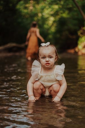 a small toddler sitting in a creek, her big sister playing behind her