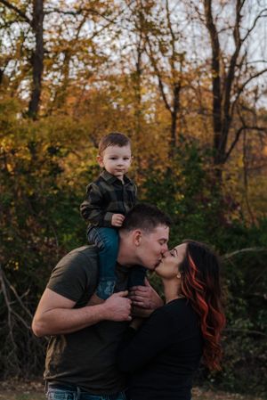 Man has a toddler boy in a flannel shirt on his shoulders, while kissing woman in black shirt.