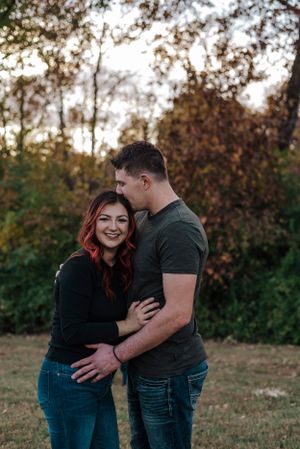 Tall man in dark tshirt kisses woman in long sleeve black shirt while she looks at camera smiling.