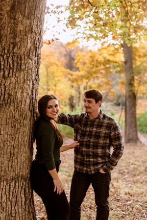 Attractive woman in dark clothing leaning up against a tree and smiling while man in flannel shirts looks at her.