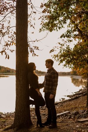 Couple standing next to lake, woman has foot propped up and leaning on a tree while man looks at her.