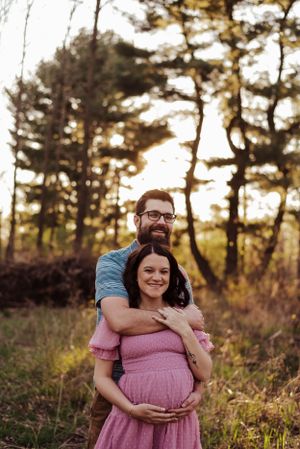 Pregnant woman standing with husband with arms wrapped around belly in sunset.