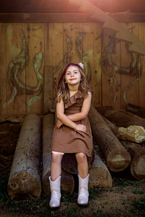 Little girl in brown dress and white boots in a barn, with sunset in the background, sitting on logs.