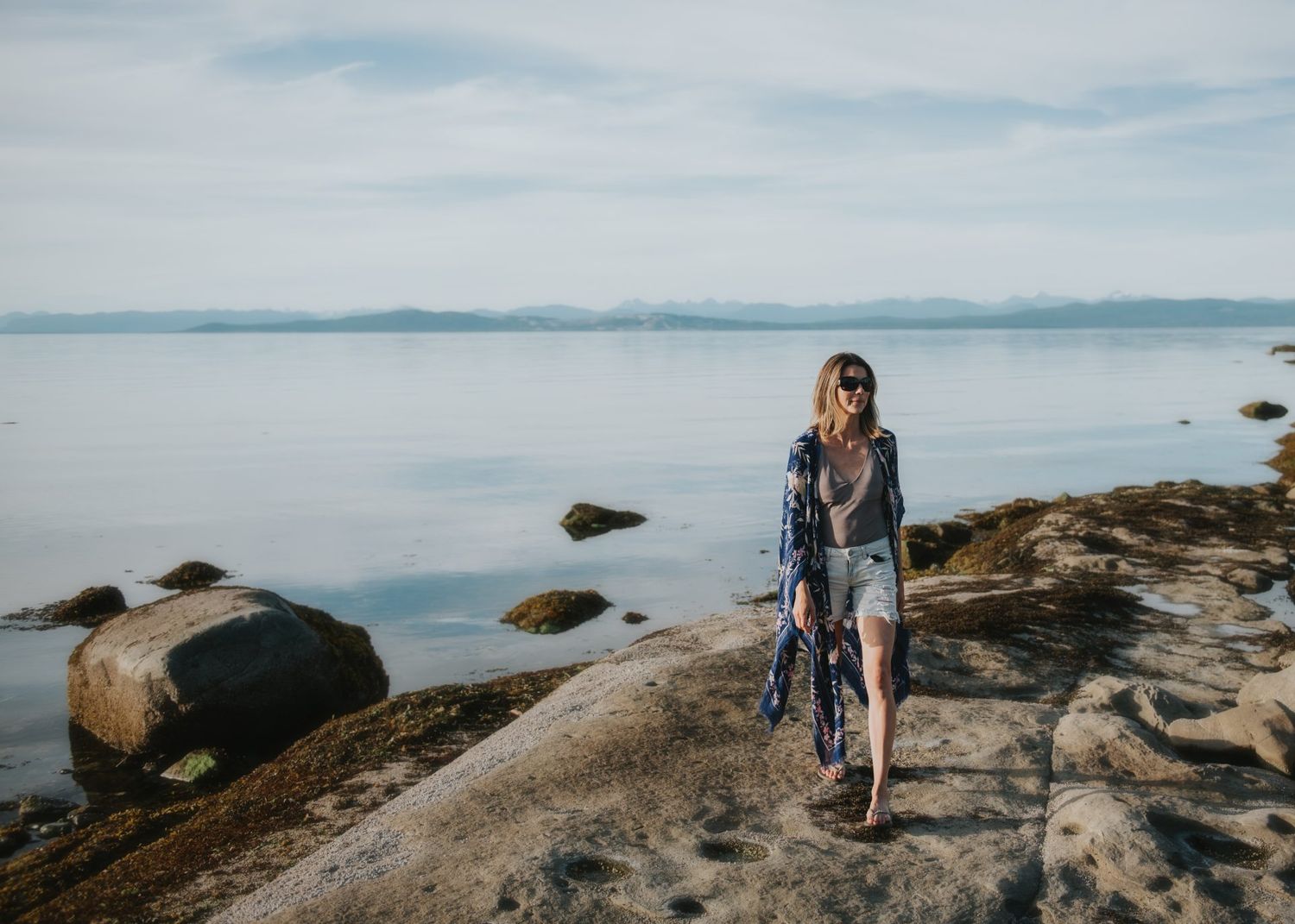 Person explores rocky coastal terrain against backdrop of calm waters and distant mountains.