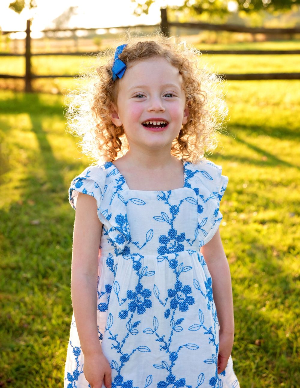 Smiling young girl with curly red hair wearing a white and blue floral dress and blue bow hair clip outdoors.