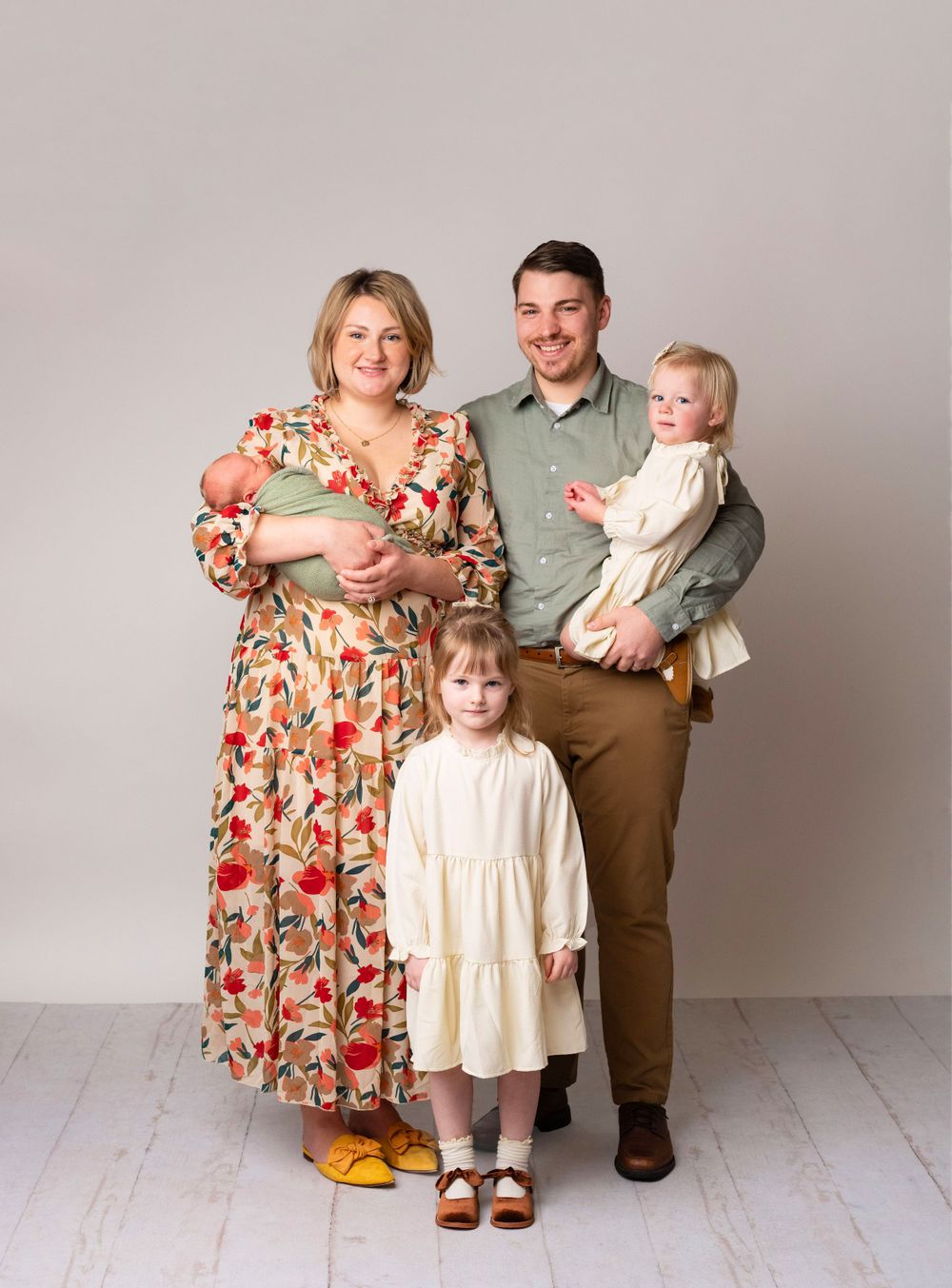 Family portrait with parents holding three young children, wearing coordinated earth-tone outfits against a neutral studio background.