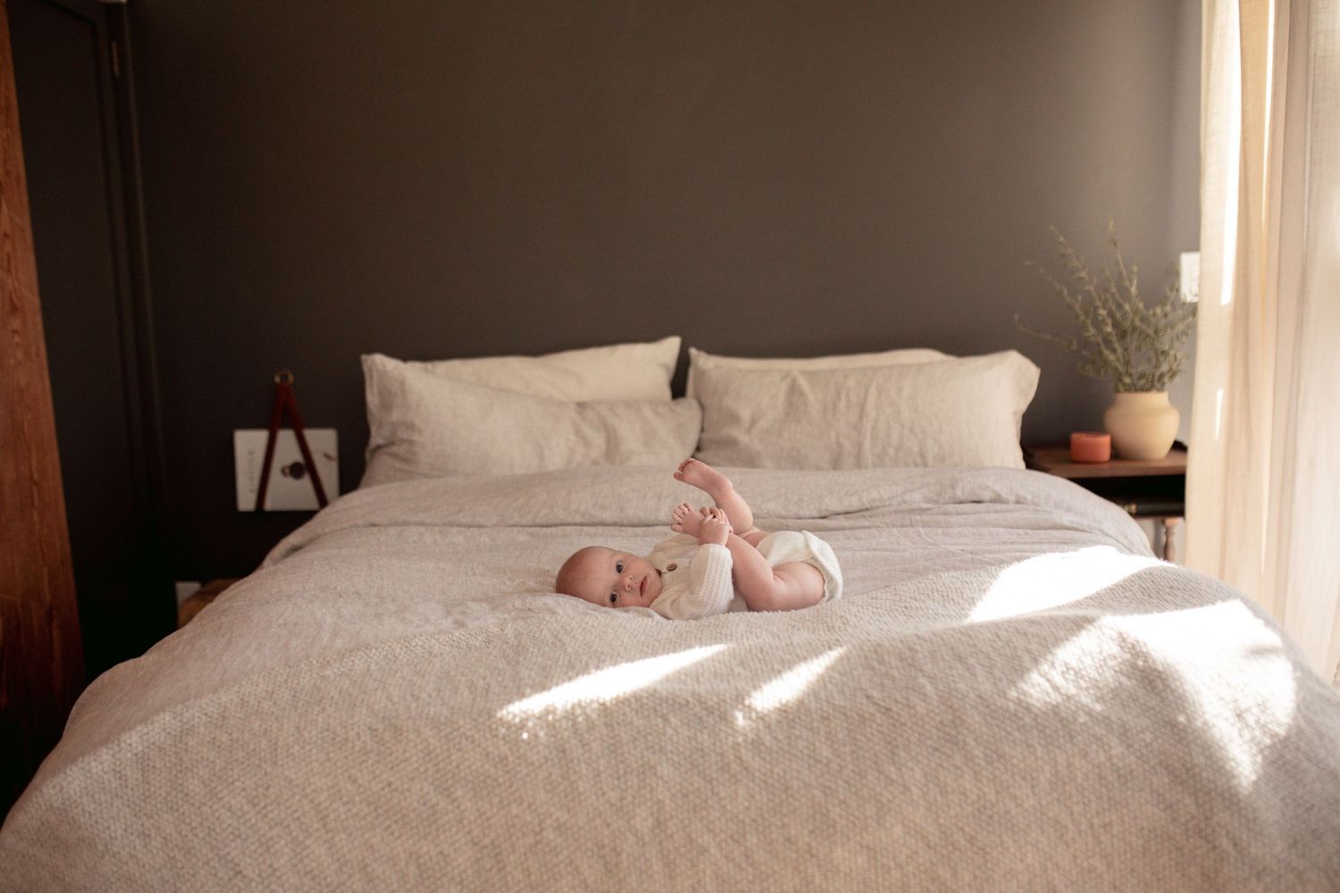 A baby resting on a white bedspread in natural sunlight streaming through a window.