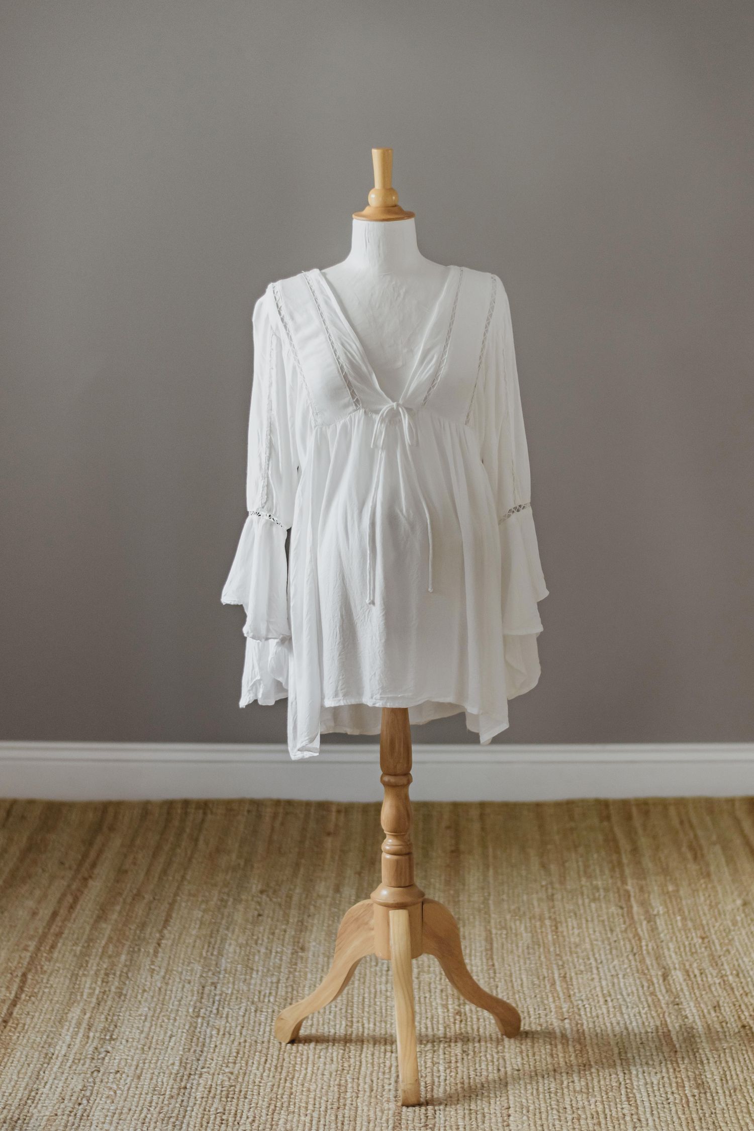 A flowy white dress with bell sleeves displayed on a wooden mannequin stand against a gray background.