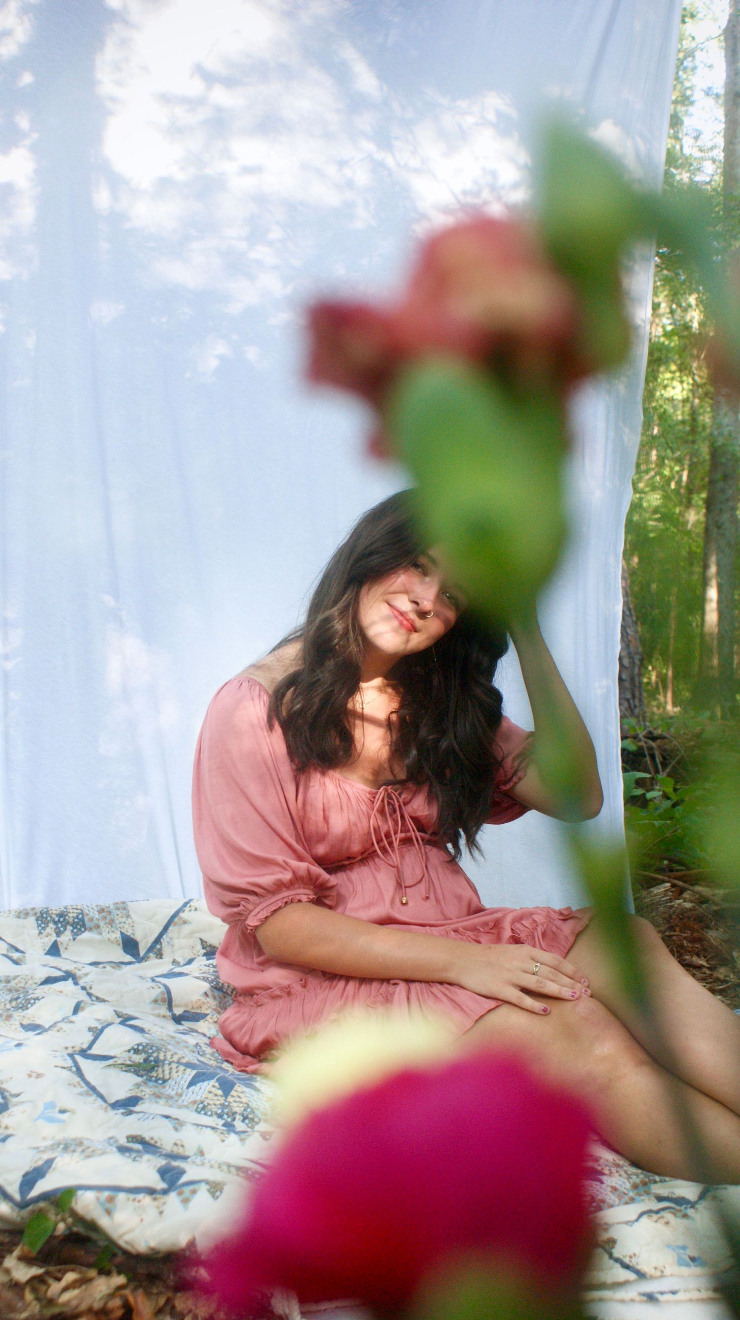 Dreamy outdoor portrait of person in pink dress reclining on white blanket with blurred floral elements in foreground.