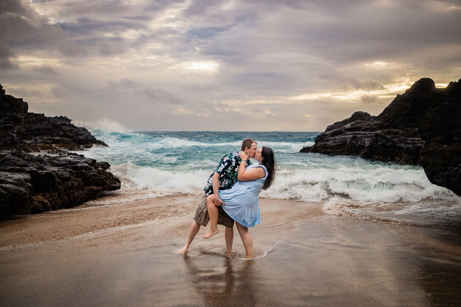 A romantic couple embraces on a beach with crashing waves and dramatic cloudy sunset sky in the background.