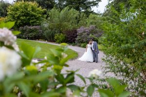 A distant wedding couple walking along a garden path surrounded by lush summer foliage.