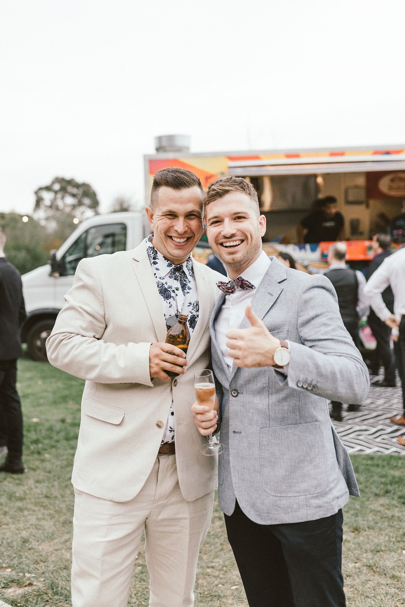 Two guests in light colored suits enjoying drinks at an outdoor wedding reception.
