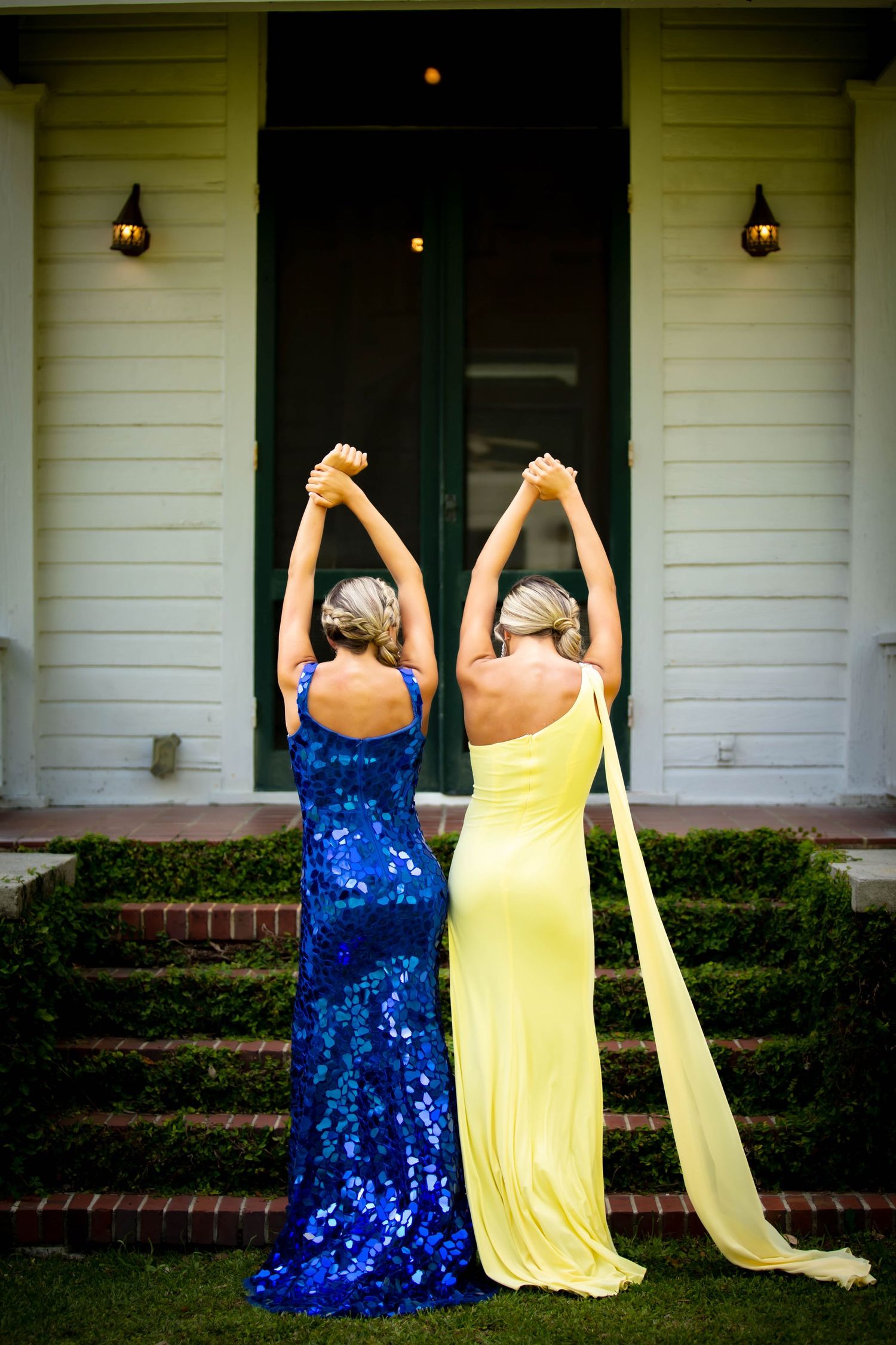 Two women in formal gowns stand back-to-back with raised arms on brick steps of a white house.