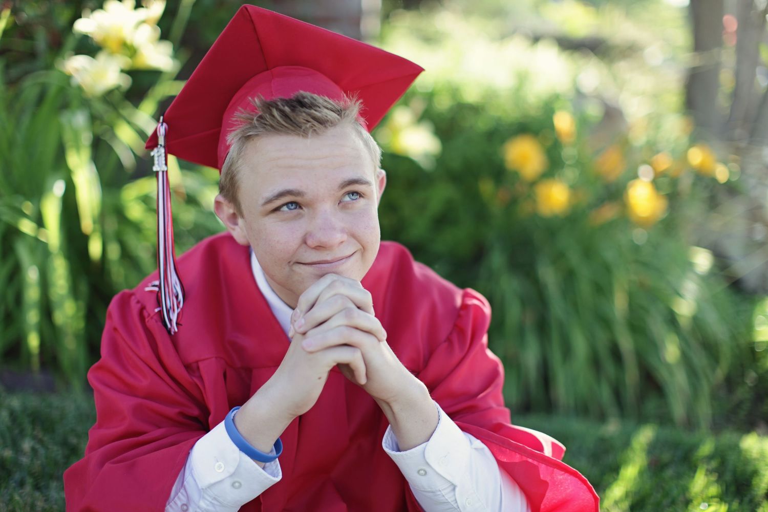 Graduate in red cap and gown poses thoughtfully in a garden setting with yellow flowers.
