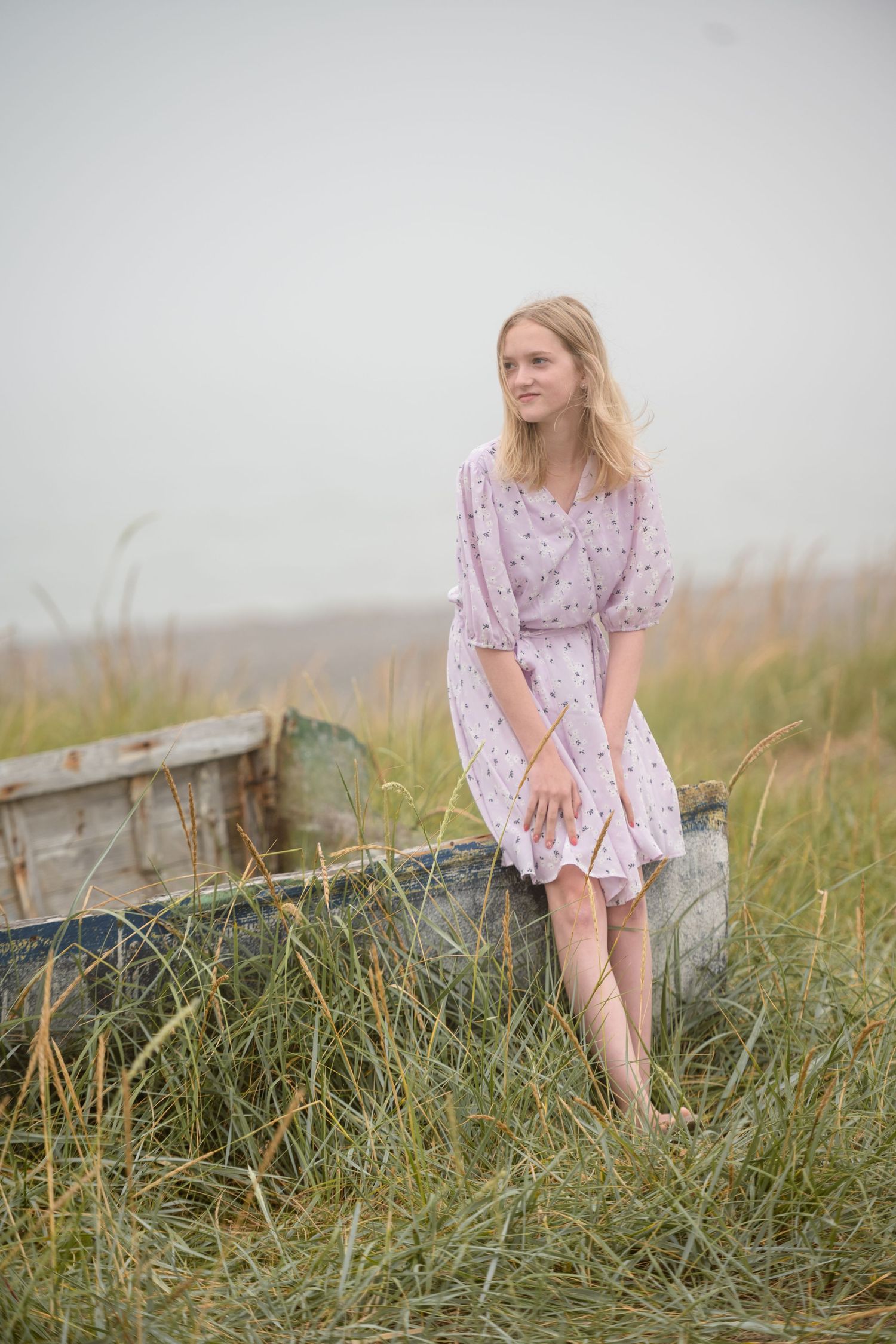 Woman in pink floral dress sitting on weathered wooden boat in tall grass.