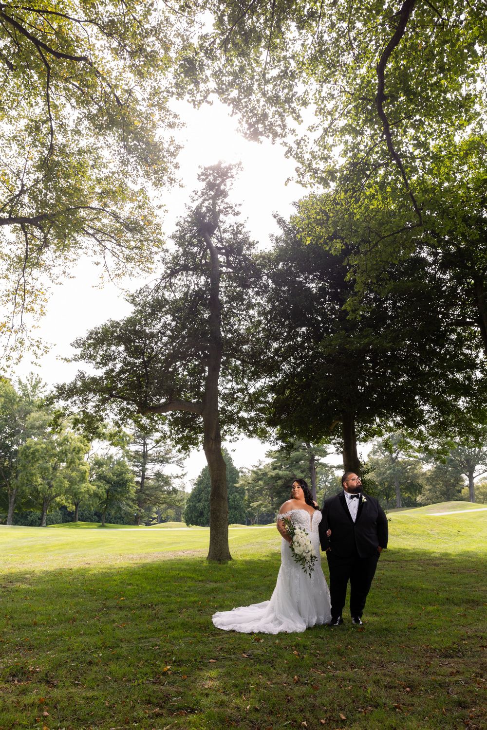 Wedding couple standing under tall trees in sunlit garden setting.
