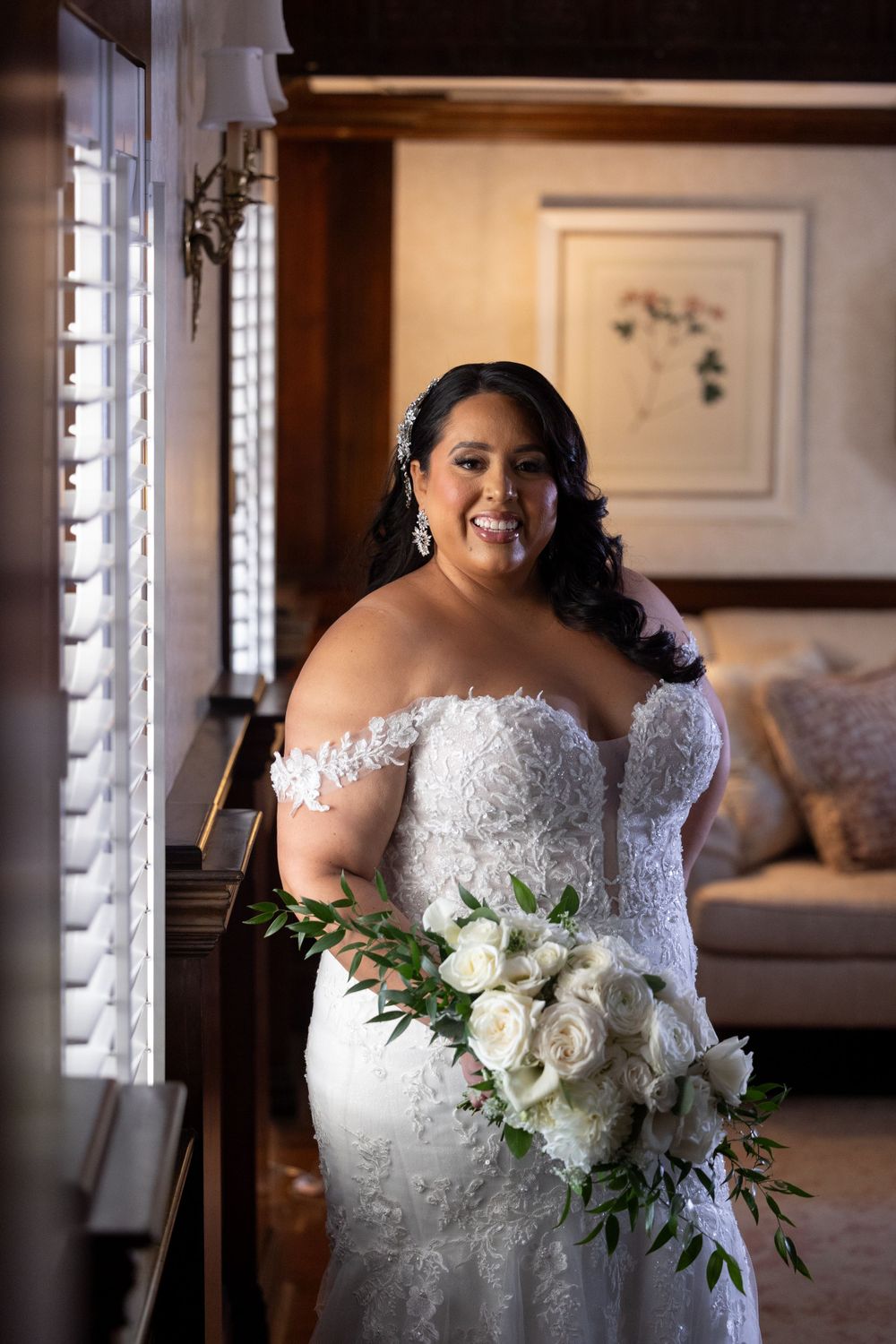 Elegant bride in lace wedding dress holding white floral bouquet by window.