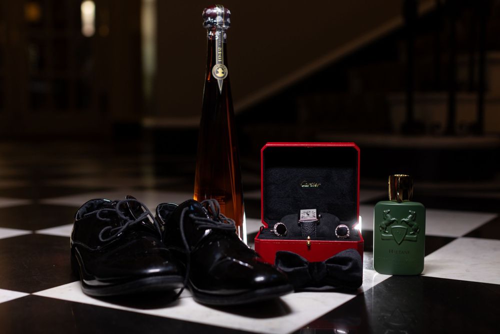 Wedding day accessories including black dress shoes, a ring box, and cologne against a dark background.