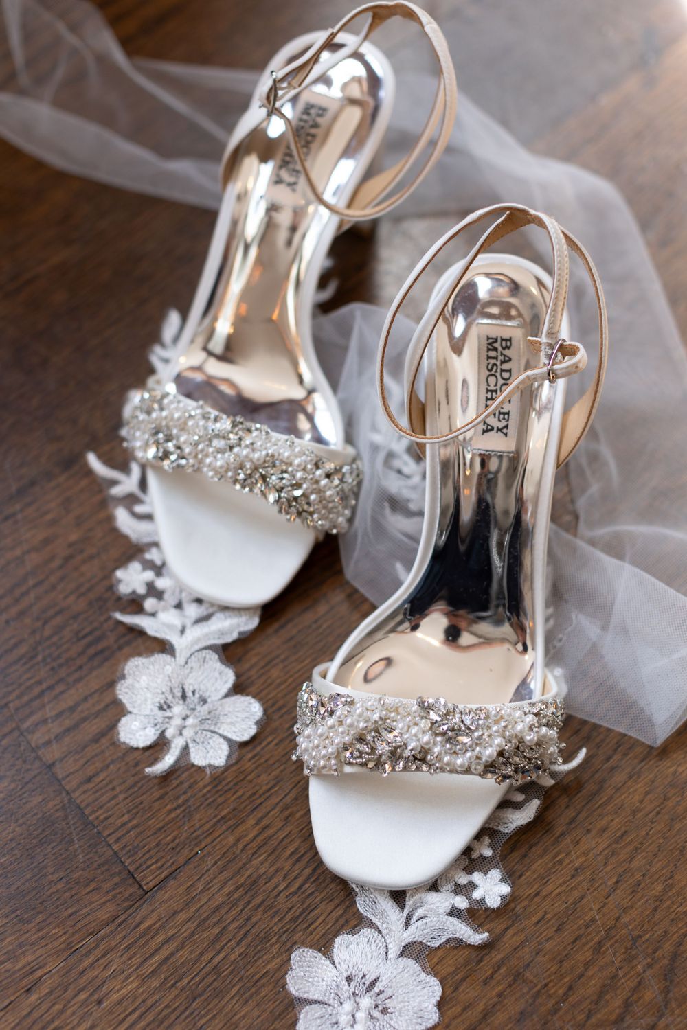 Crystal-embellished white bridal heels with delicate lace floral accents on a wooden surface.