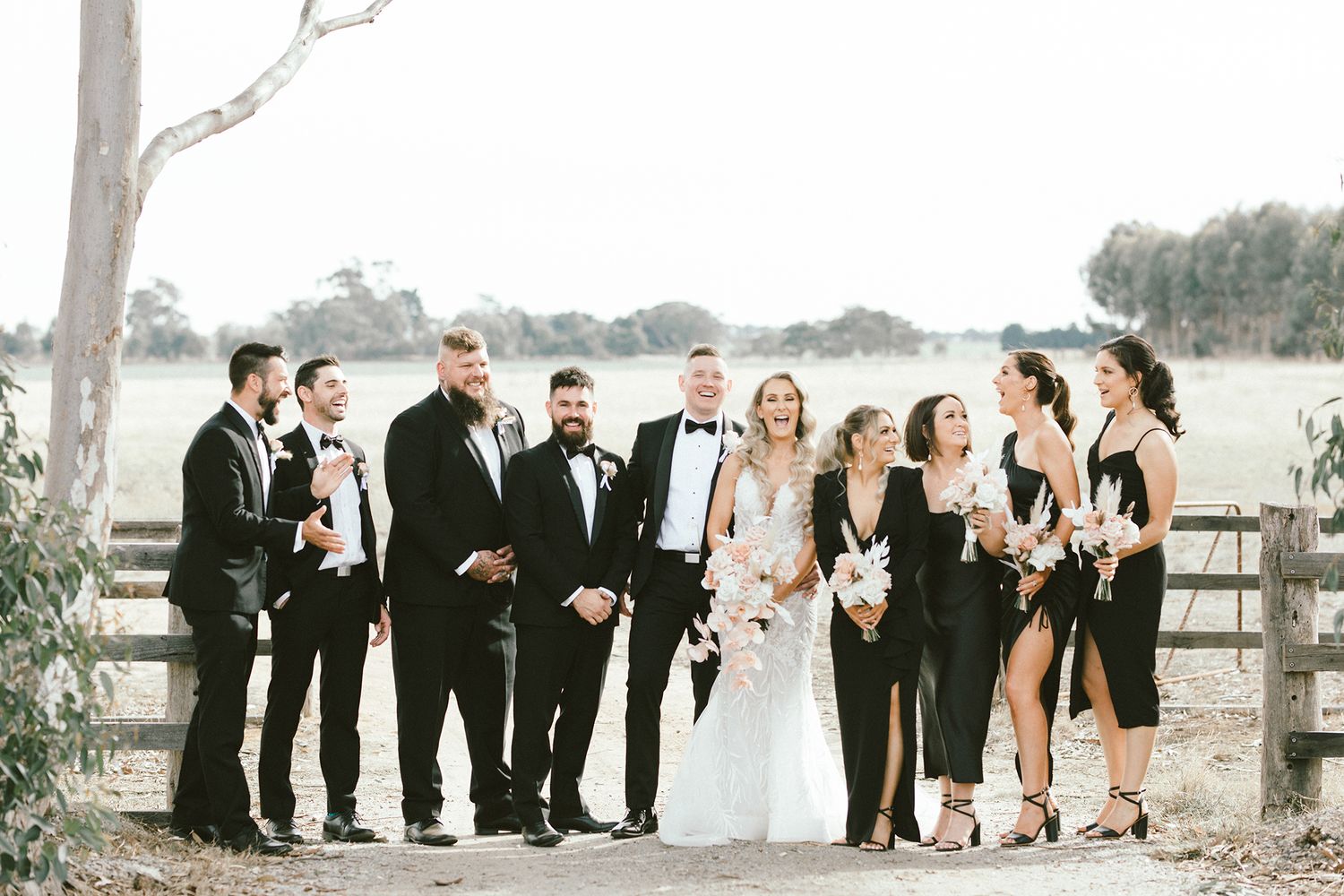 Candid moment of wedding party laughing together by wooden fence and trees.