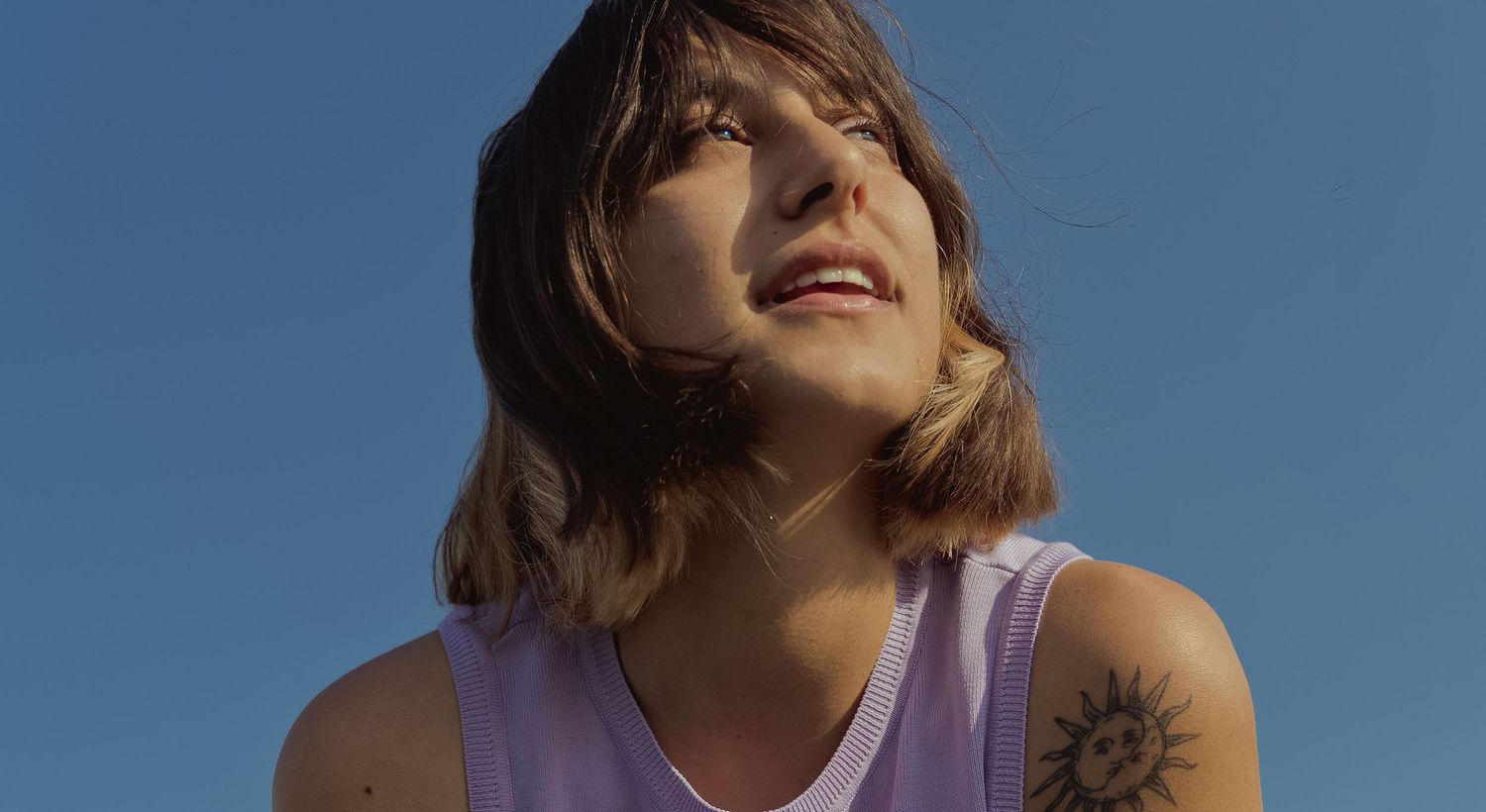 A candid portrait against bright blue sky with a visible arm tattoo.