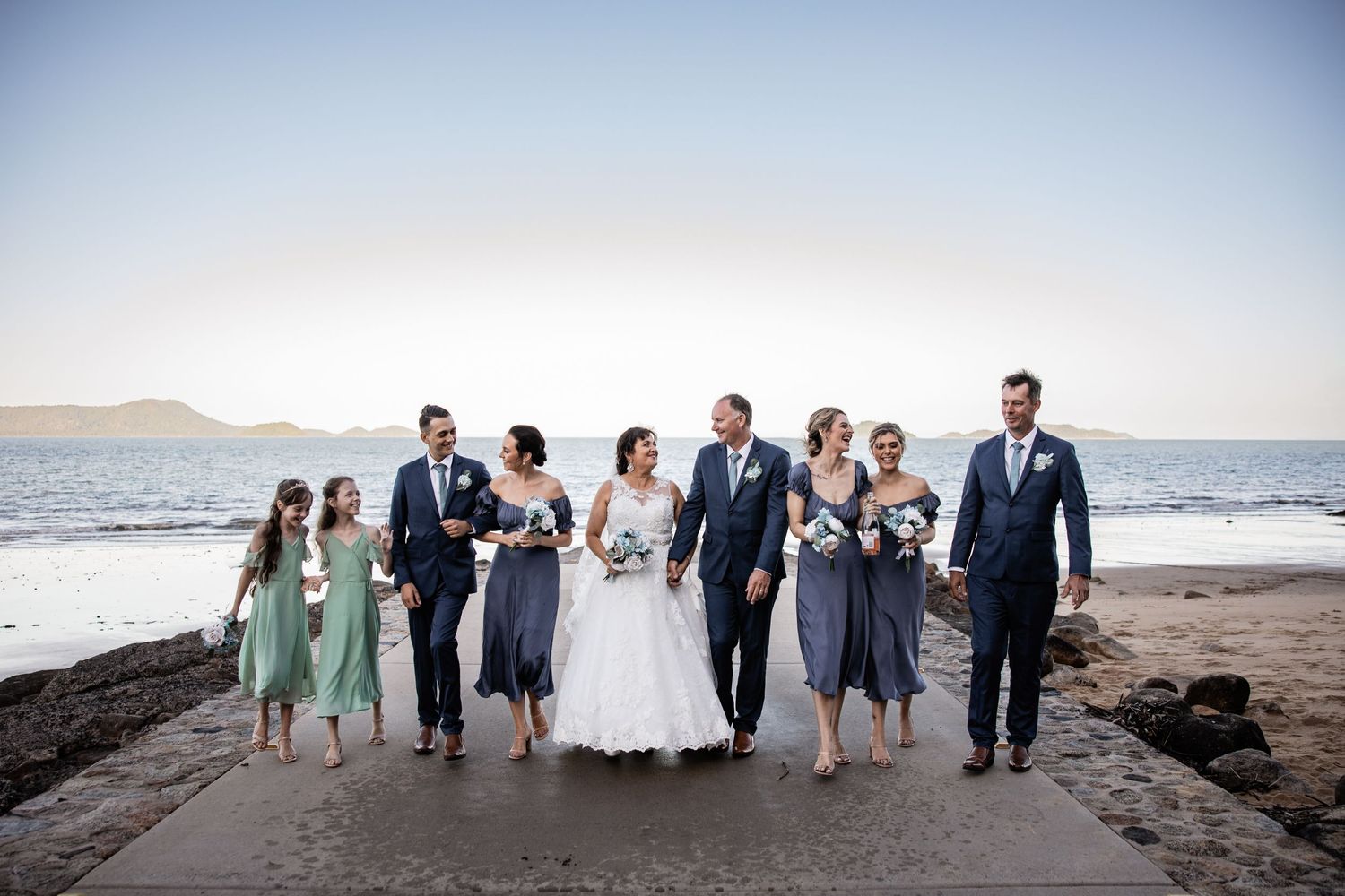 Wedding party walks along a rocky beach shoreline with ocean and mountains in the background.