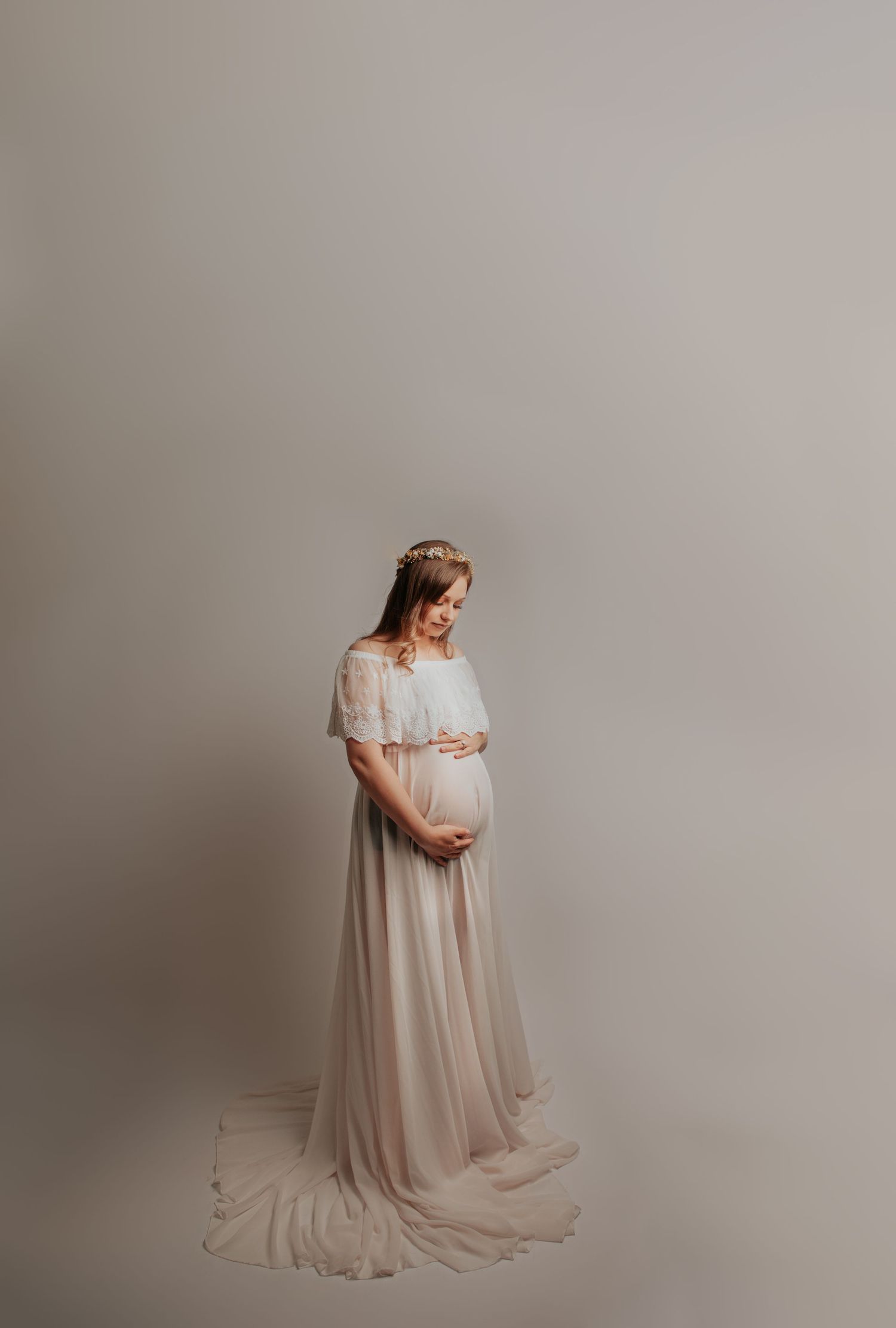 Expectant mother poses in flowing white gown against neutral studio backdrop.