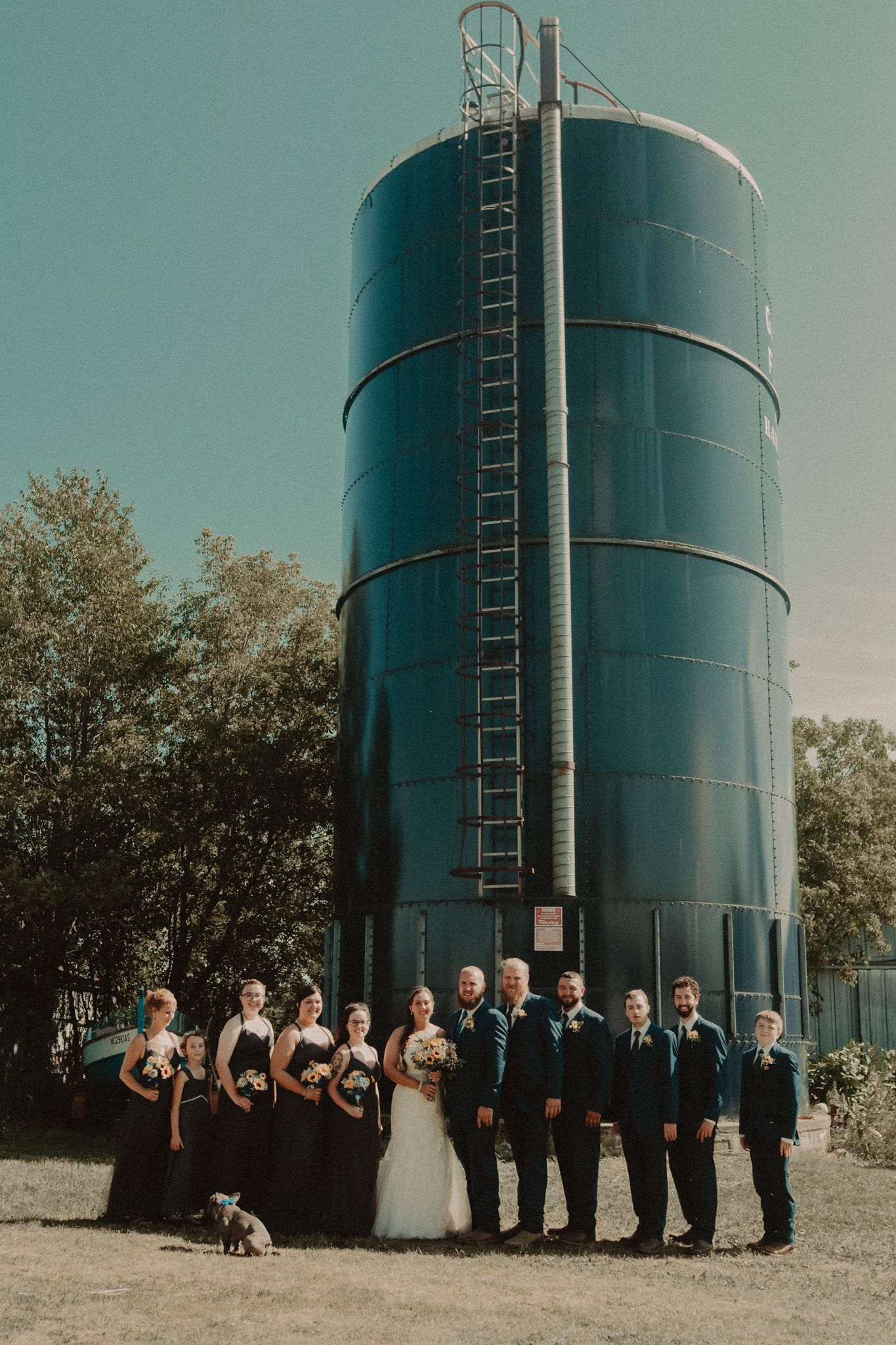 A wedding party poses in front of a large blue silo with bridesmaids in dark dresses.