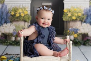 Baby girl in dark blue, sitting and smiling on a tiny bed. The background is yellow and blue spring flower bunches.
