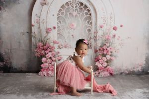 A baby girl sitting on a tiny white bed, smiling and wearing a pink dress. The background is a pretty white arch with pink flowers