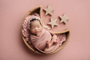 A newborn baby girl sleeping in a wooden moon prop on a light pink background with a few wooden stars nearby.