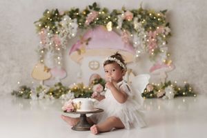 Baby girl with a first birthday cake, wearing a white dress and white fairy wings. The background is a mushroom house with cascading flowers