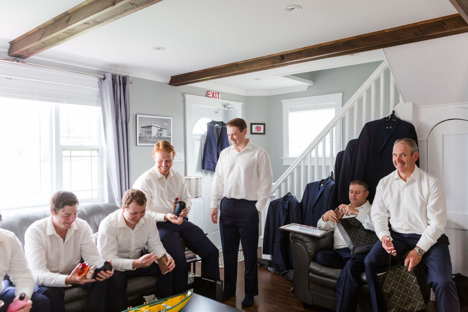 Groomsmen getting ready and sharing laughs in bright white room with wooden beams.
