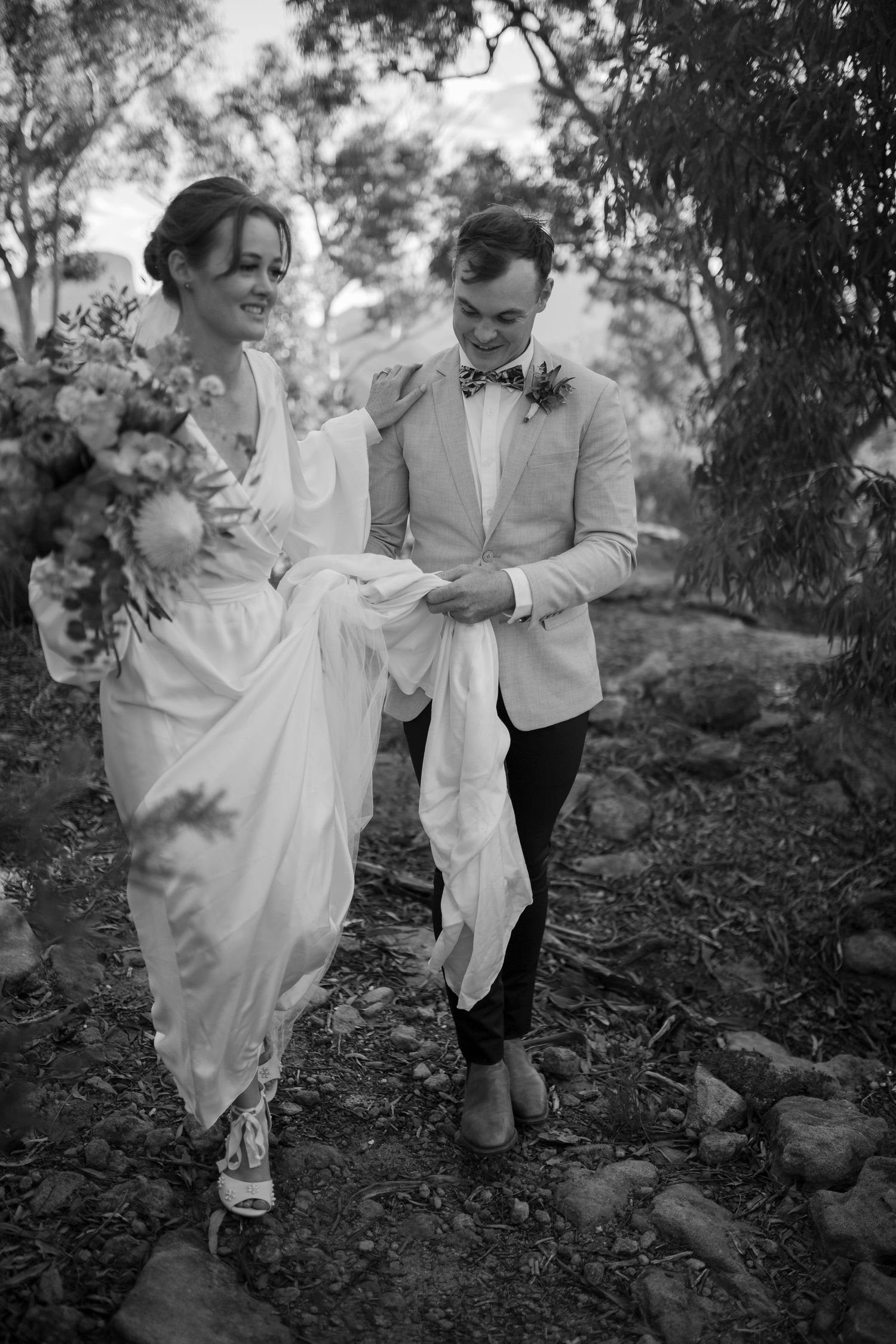 Black and white photo of couple walking together on rustic path in formal attire.